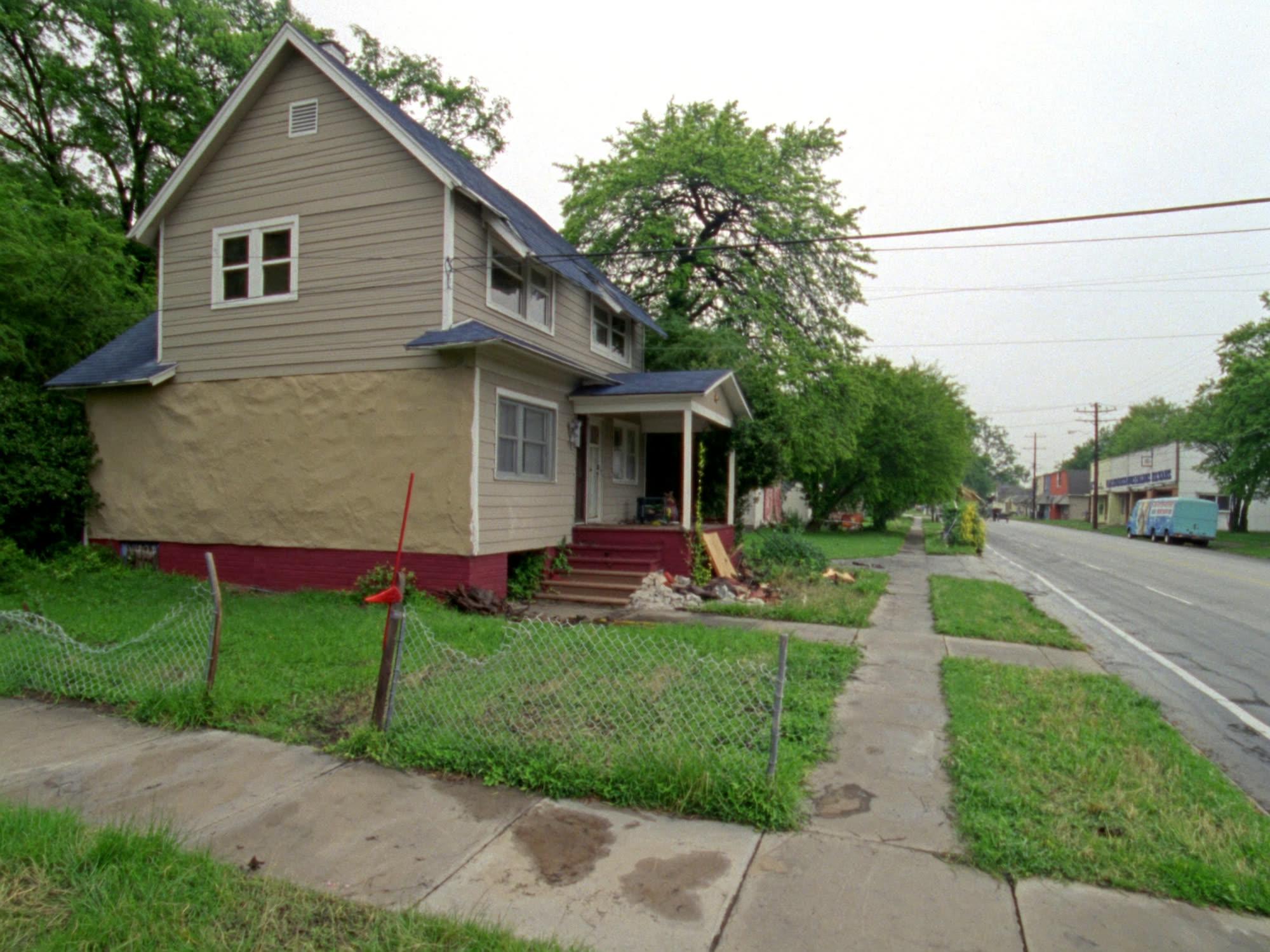 A tired two-story house with faded paint, an overgrown yard and a collapsing chain-link fence.