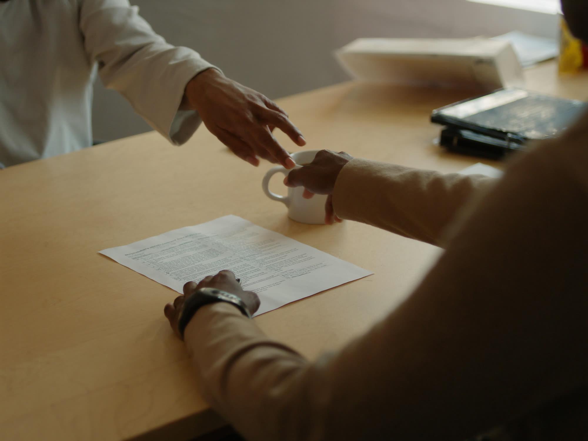 Two people seated across a table, hands resting on a housing application document beside a coffee mug in warm window light.